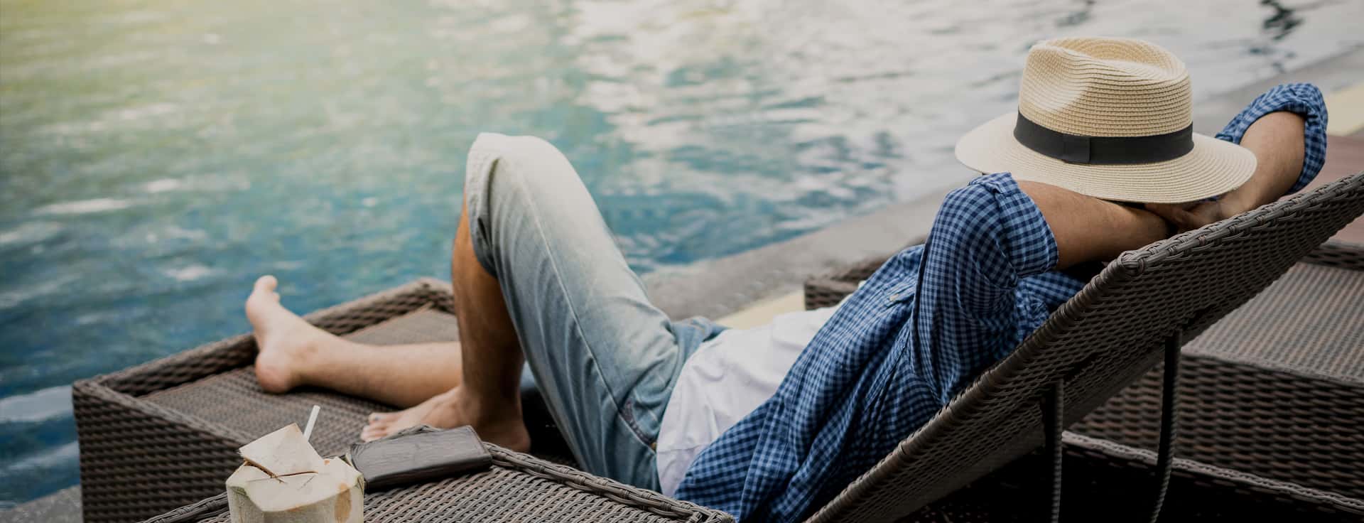 Wide darkened shot of a happy man with a hat who is relaxing on a sunbad next to a pool filled with sustainably desalinated water. The man has no bad conscience anymore as the water is purified in a sustainable way.