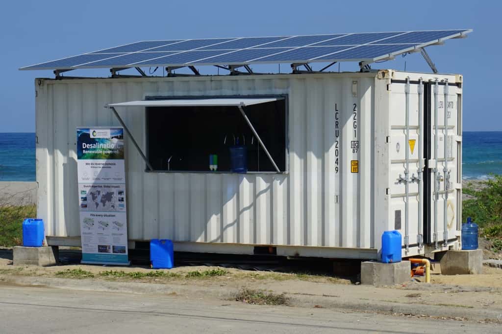 Our water kiosk in Madagaskar in action, with a poster about general information and instructions in the front The container's side is opened and the faucet placed in such a way that people can come and collect their drinking water. Water containers are provided to the local community to ensure clean drinking water during tranportation. There is a beach and the ocean on the background, illustrating the source of the drinking water.