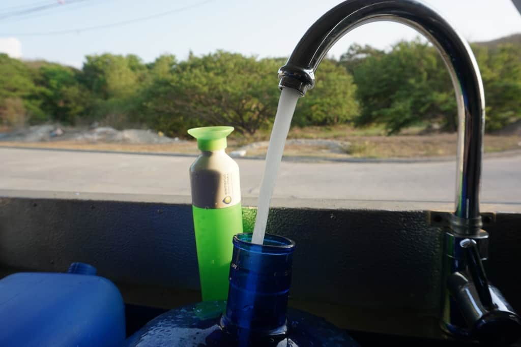 A "Dopper" bottle placed next to a faucet, with the inner side of the desalination container in the beackground. There is drinking water flowing out of the faucet, which is the final product of the reverse osmosis desalination system and was produced sustainably without negative effects on the environment or communities.