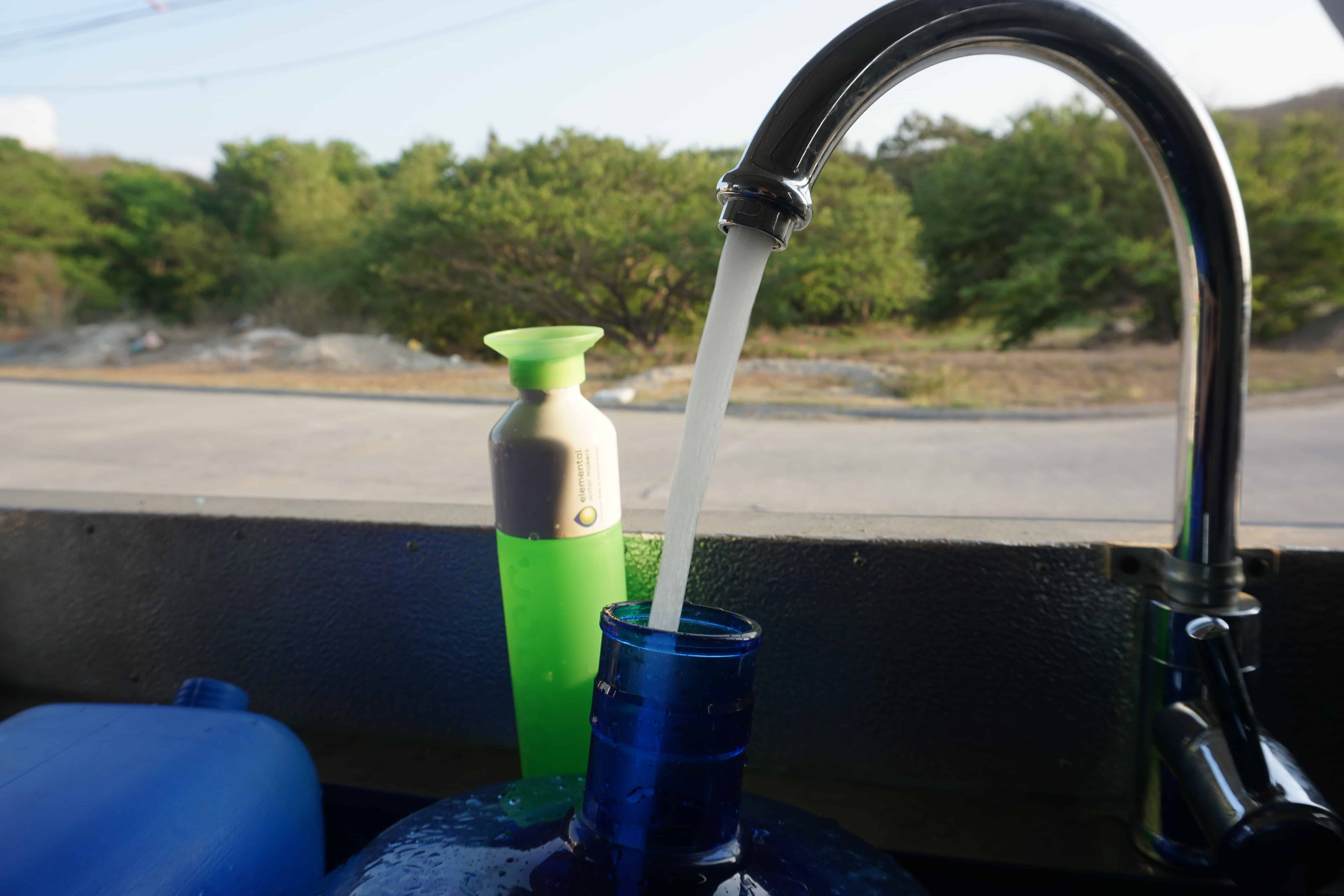 A "Dopper" bottle placed next to a faucet, with the inner side of the desalination container in the beackground. There is drinking water flowing out of the faucet, which is the final product of the reverse osmosis desalination system and was produced sustainably without negative effects on the environment or communities.