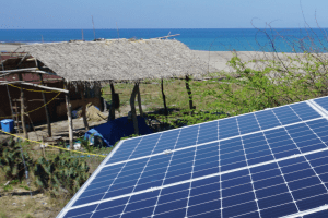 Solar panels that are providing the necessary power to run the desalination system. The generated energy is partly stored in a battery (with a long life-span), which ensures water supply in periodes with shortages in sunlight. In the background there is a small fishing hut and a beach.